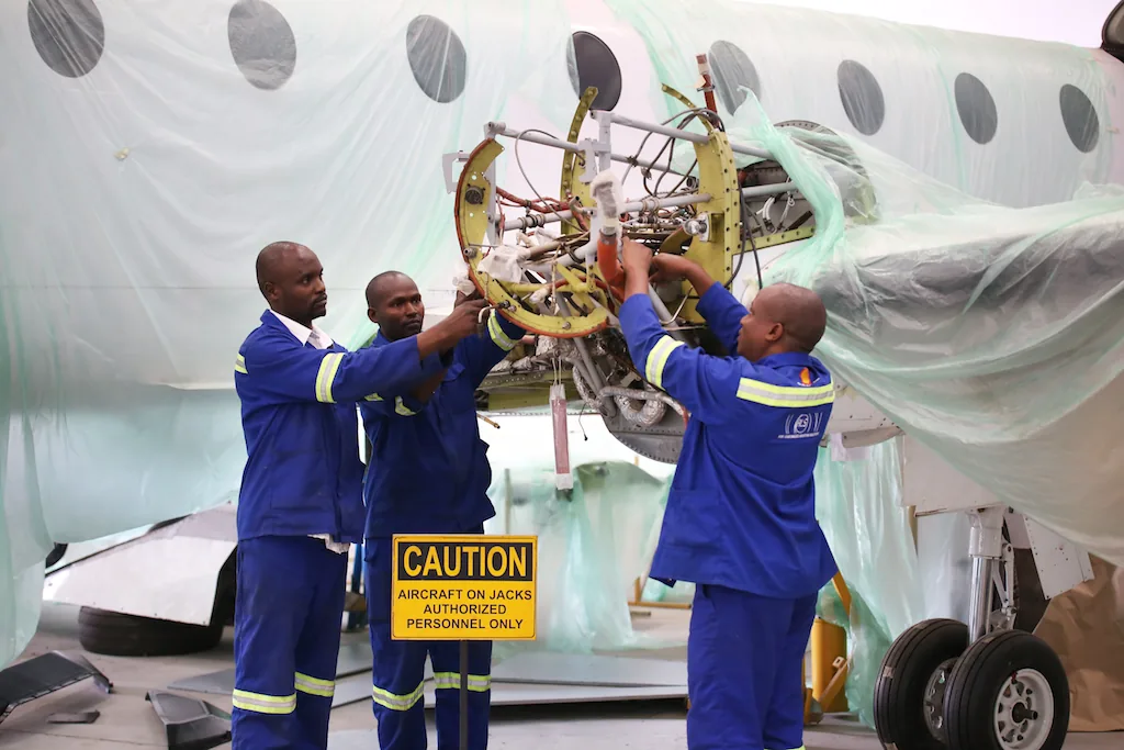 Three ALS engineers in blue overalls lifting a component beside an aircraft in the hangar.